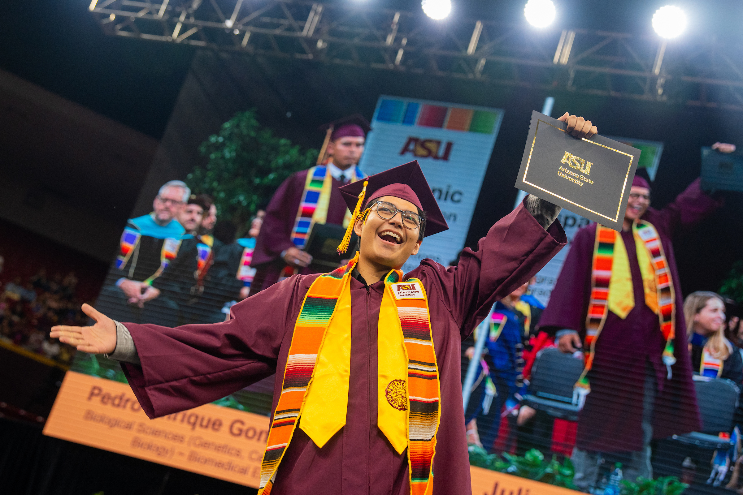 Male, Hispanic Student, wearing ASU maroon cap and gown, accompanied with his serape, ASU Stole of gratitude, and holding his black with gold lettering ASU Certificate folder. 